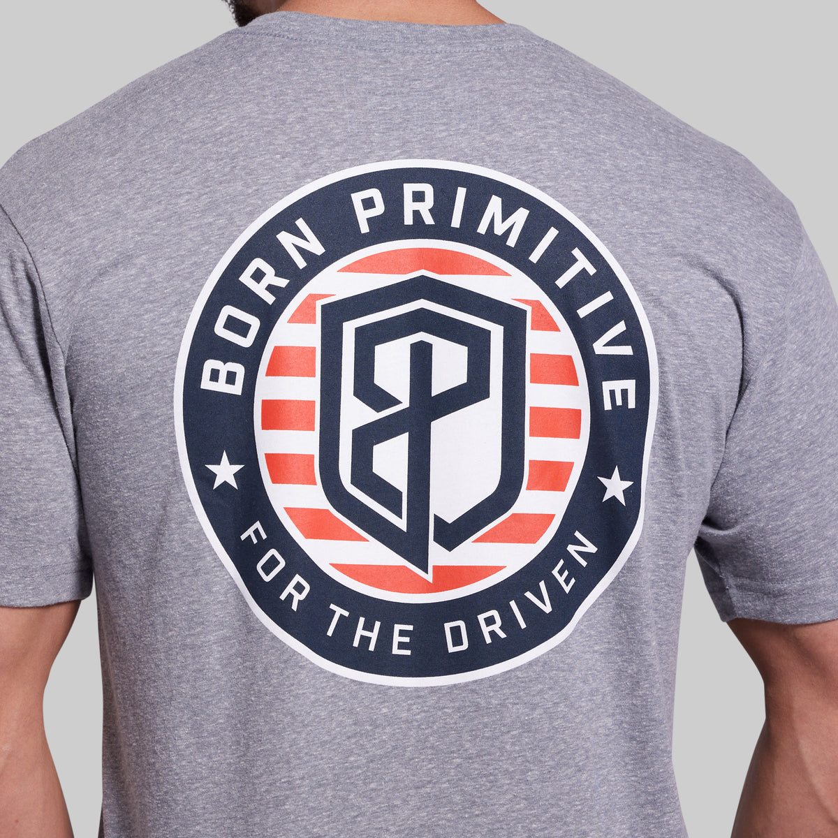 A close-up of a heather gray t-shirt showing the back with a large circular graphic. The graphic features the text 'BORN PRIMITIVE FOR THE DRIVEN' in white around a shield logo with red and white stripes and a navy blue 'BP' monogram in the center.