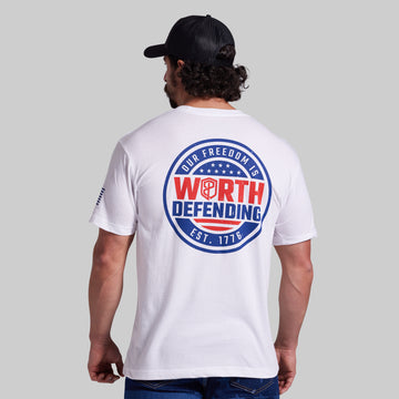 A man viewed from behind wearing a white t-shirt with a large patriotic back graphic that reads 'OUR FREEDOM IS WORTH DEFENDING EST. 1776' in red, white, and blue, paired with a black baseball cap and blue jeans, against a solid gray background.