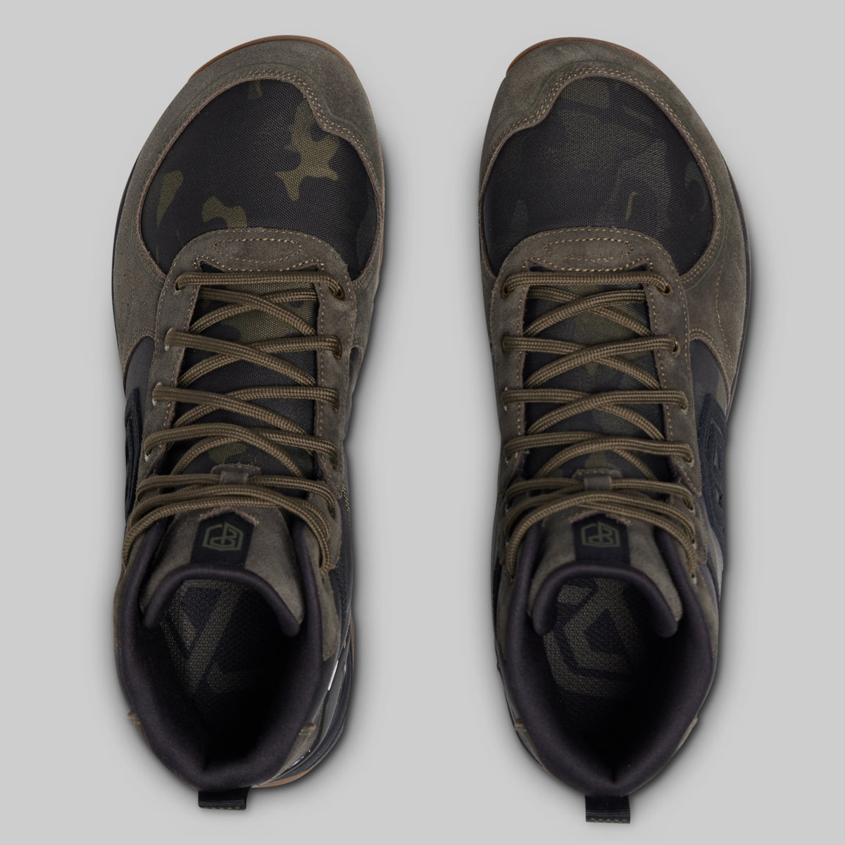 A pair of high-top outdoor boots shown from a top-down view, featuring a dark camouflage pattern on the toe and side panels, olive green suede overlays, and matching olive laces, set against a solid gray background.
