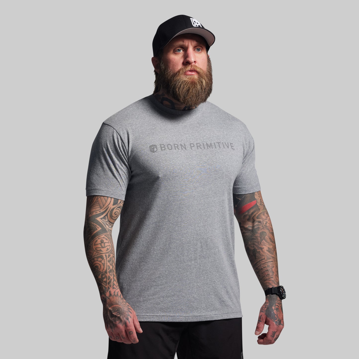 Man wearing a gray 'Born Primitive' t-shirt and black cap on a light gray background