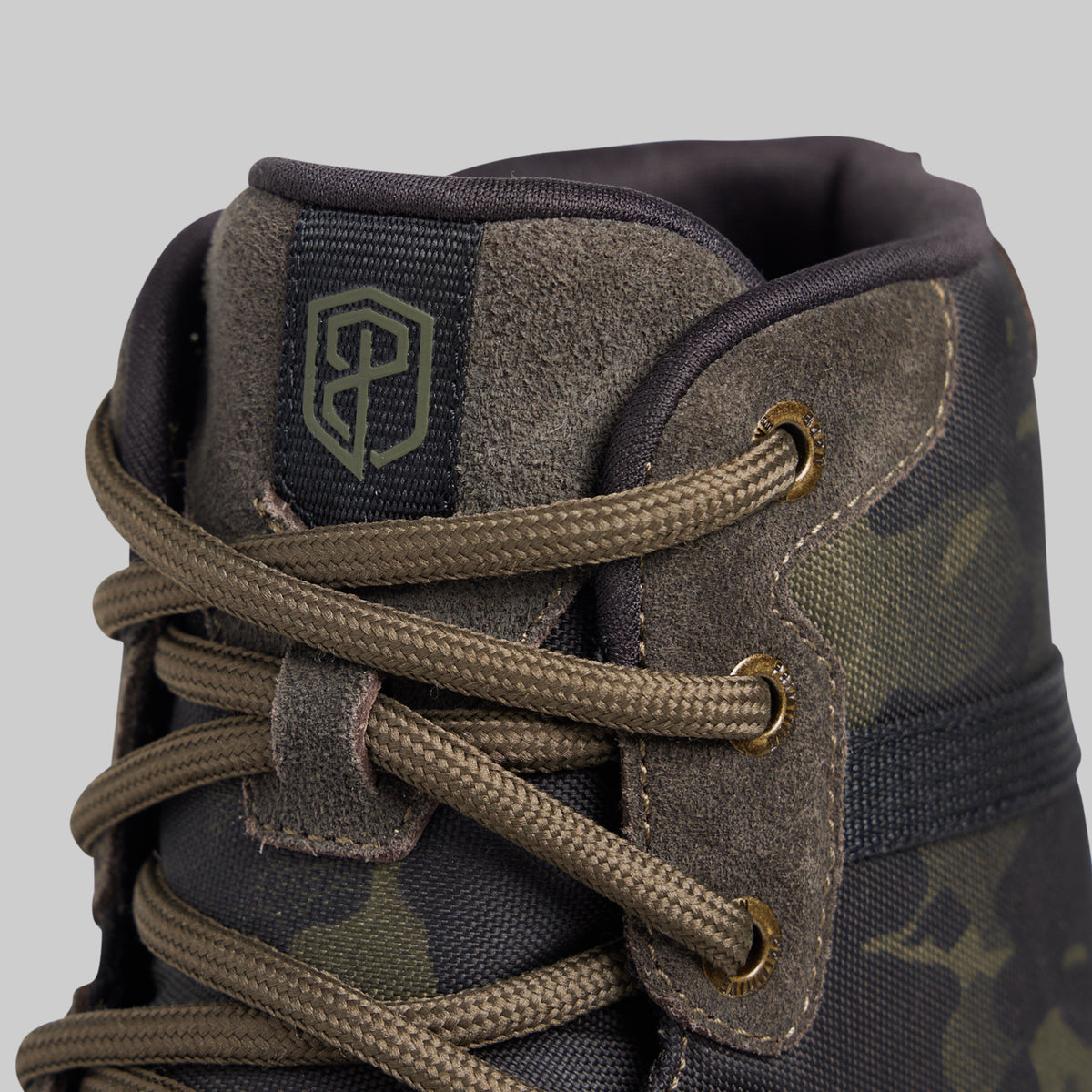 Close-up of a high-top lace-up boot in dark olive green and black camouflage fabric, featuring brown laces threaded through brass eyelets and a dark green embroidered shield logo on the tongue patch.