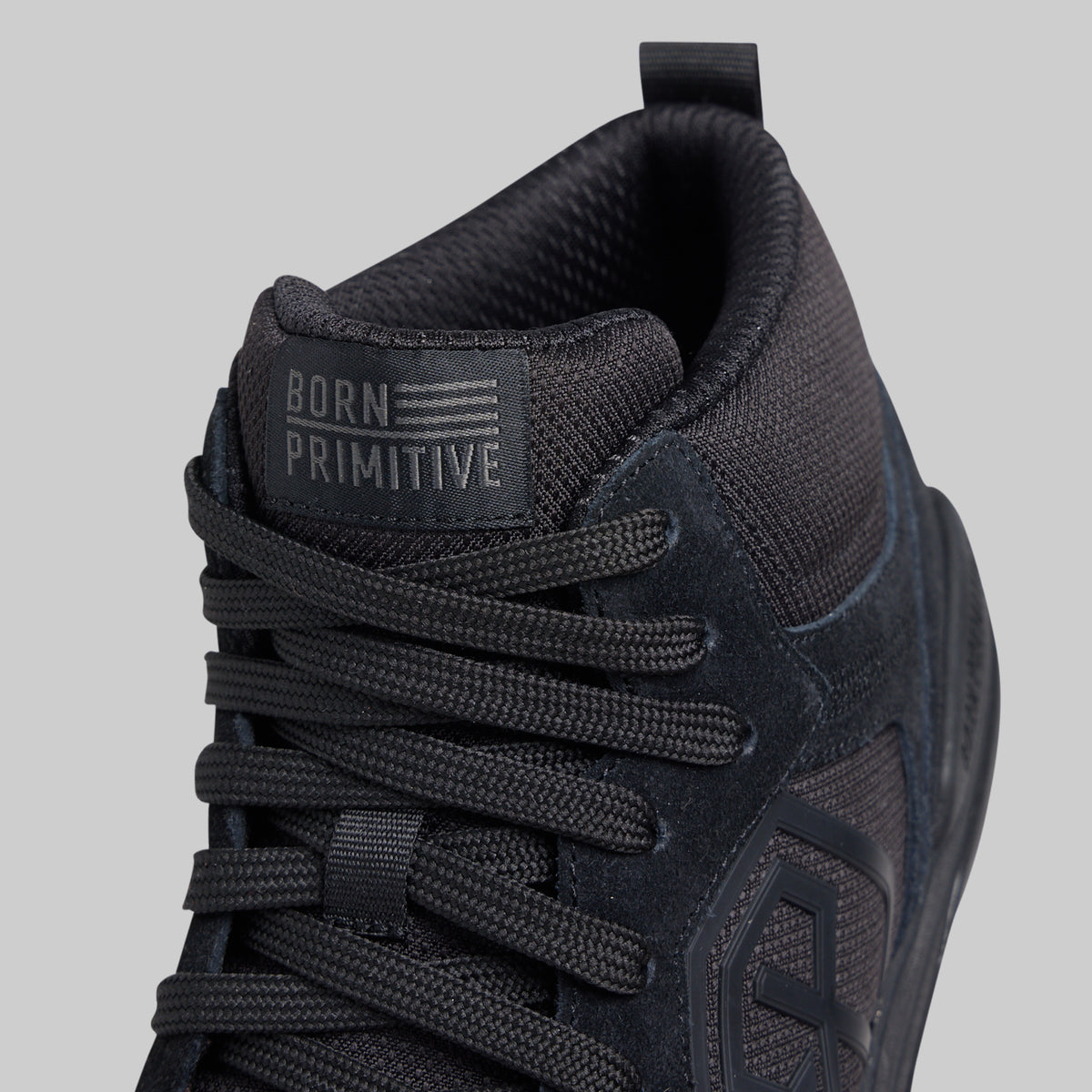 Close-up of a black mid-top sneaker by Born Primitive, showing the textured mesh and suede upper, black laces, and the brand's tongue label.