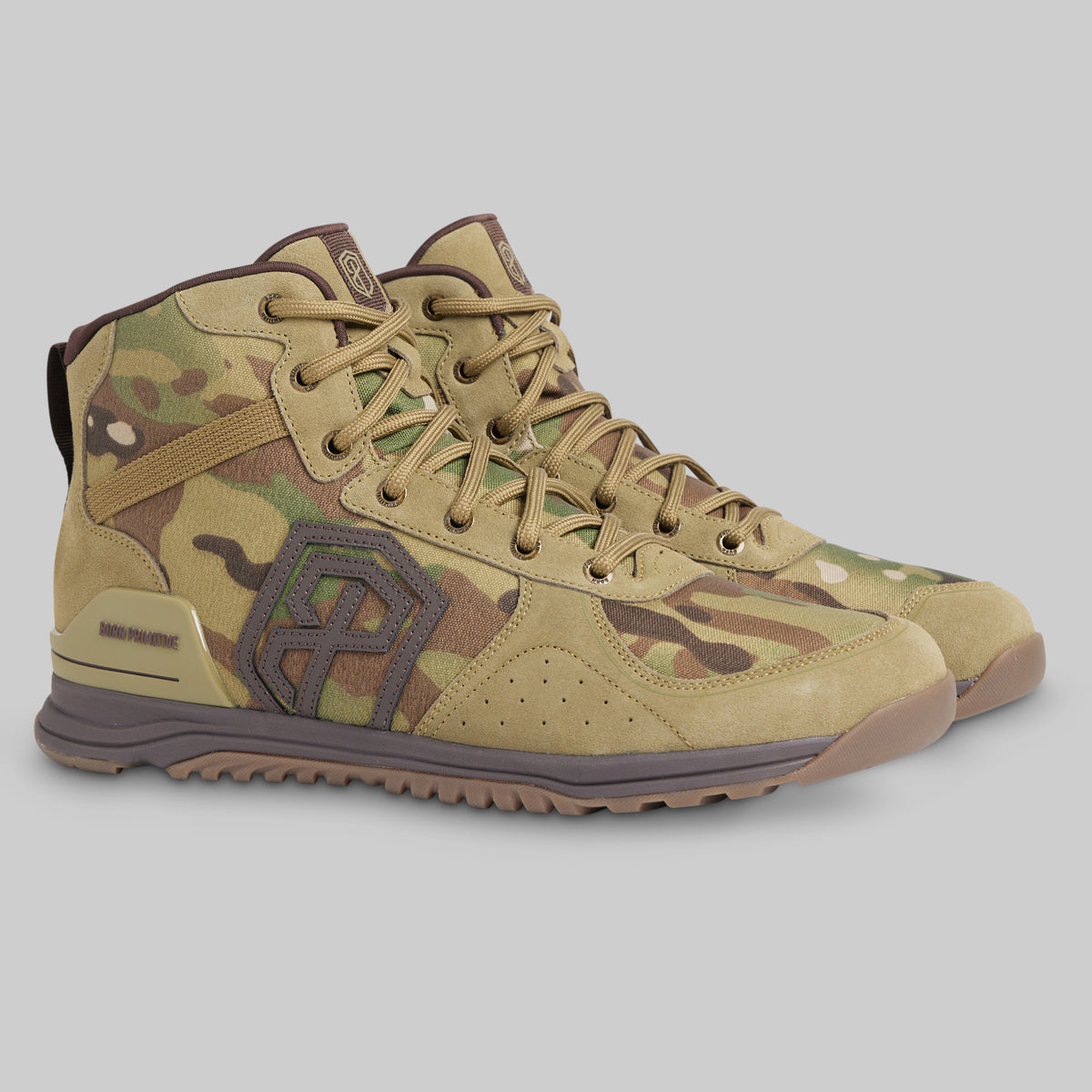 A pair of mid-top tactical boots in a tan and brown camouflage pattern, featuring a large geometric logo on the side, tan laces, and a rugged sole, set against a solid gray background.