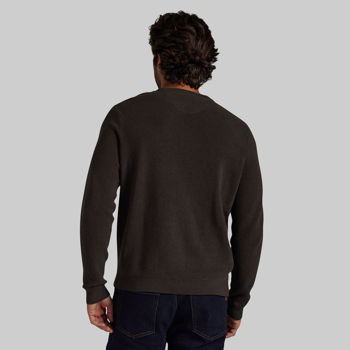 A man viewed from behind wearing a dark brown crewneck sweater and dark blue jeans, set against a solid light gray background.