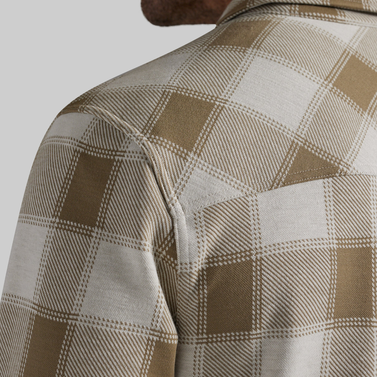 Close-up of a person wearing a plaid overshirt in beige and off-white, showing the shoulder, upper back, and part of the neck against a light gray background.