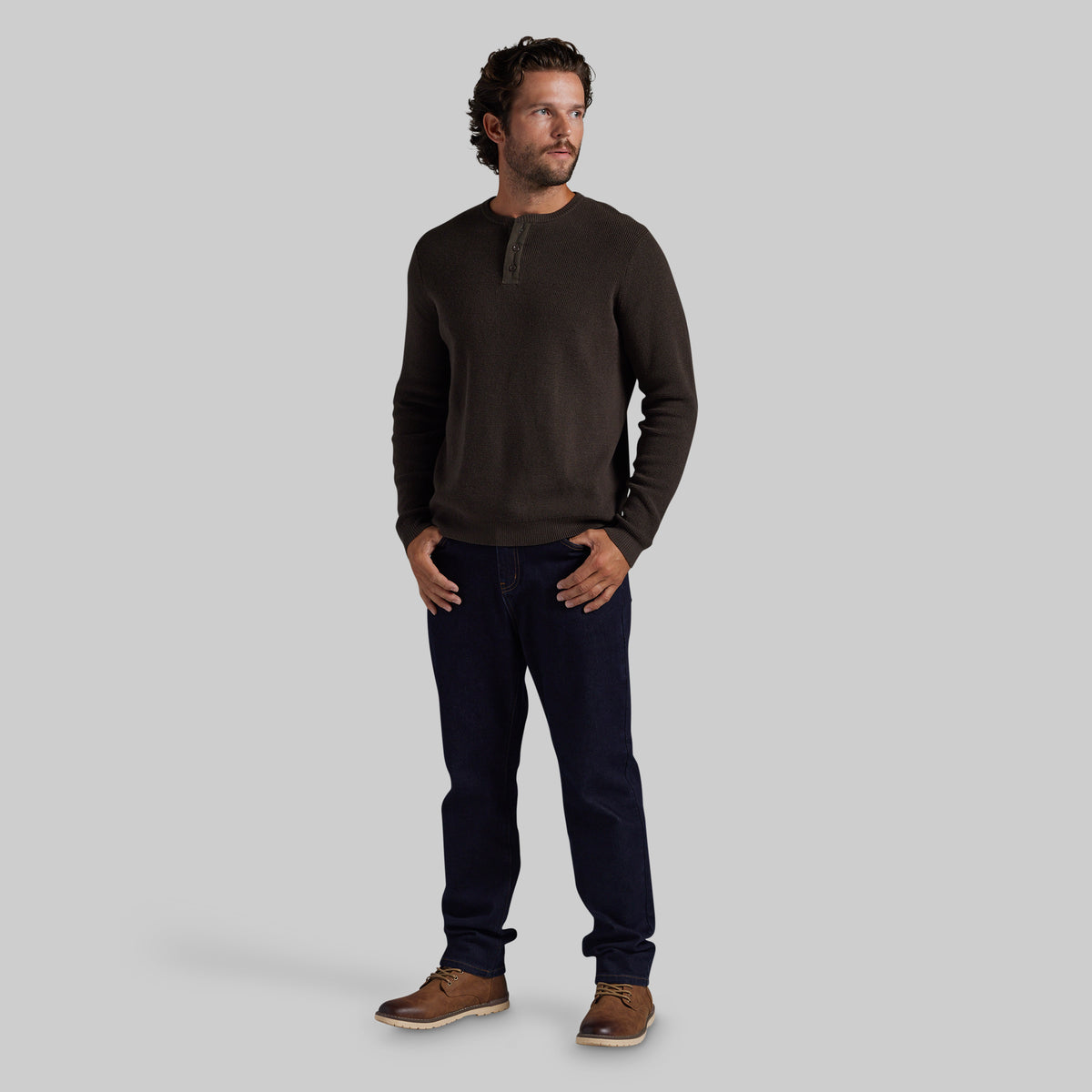 A man wearing a dark brown long-sleeve henley shirt, dark navy blue jeans, and brown leather shoes, standing against a solid gray studio background.