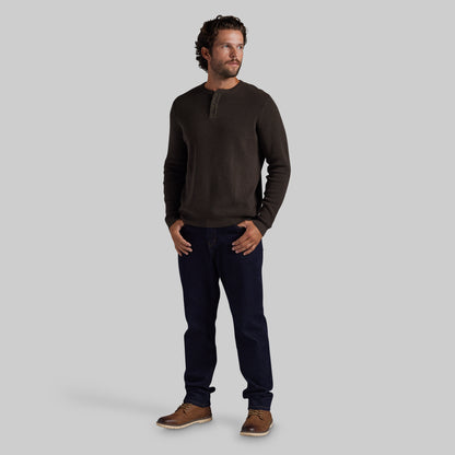 A man wearing a dark brown long-sleeve henley shirt, dark navy blue jeans, and brown leather shoes, standing against a solid gray studio background.