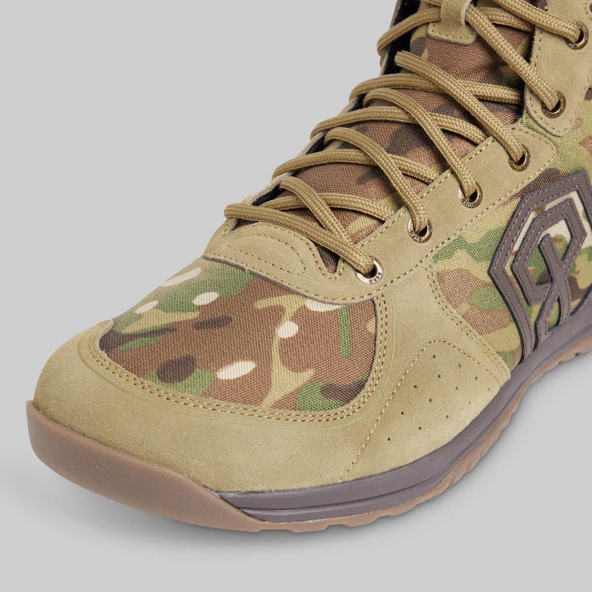 A close-up of a tactical boot featuring a tan suede and camouflage upper with matching laces, a brown sole, and a dark brown logo on the side, set against a solid gray background.