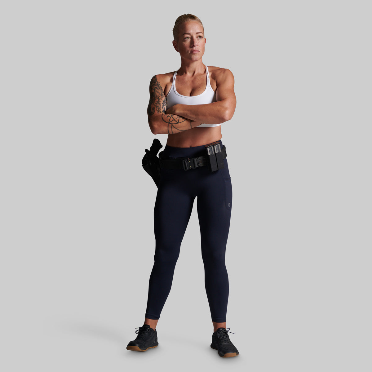 A woman stands against a light gray studio background wearing dark navy leggings, a white sports bra, and black athletic shoes. She has her arms crossed and a black tactical belt around her waist.