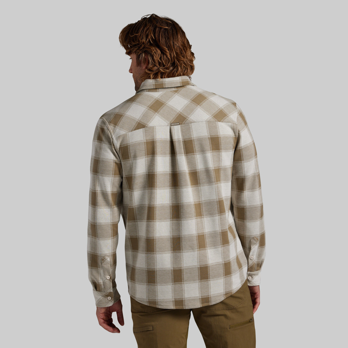 A man viewed from behind wearing a long-sleeve plaid button-up shirt in beige and white, paired with brown pants, against a solid gray background.