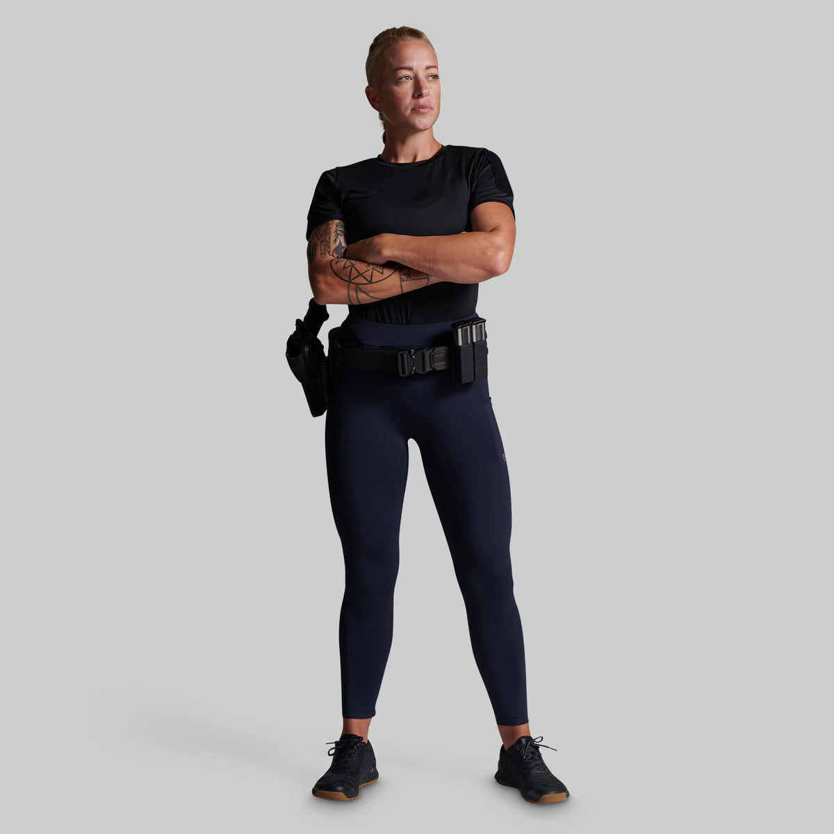 A woman stands against a light gray studio background wearing a fitted black short-sleeve top, dark navy leggings, a black utility belt with pouches, and black sneakers.