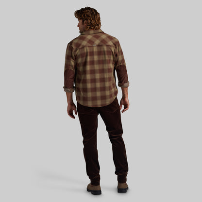 A man shown from the back wearing a brown and tan plaid flannel shirt, dark brown corduroy pants, and brown boots, set against a solid light gray background.