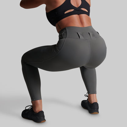 A model from behind wearing dark gray high-waisted athletic leggings, a black sports bra, and black sneakers, performing a deep squat against a solid gray studio background.