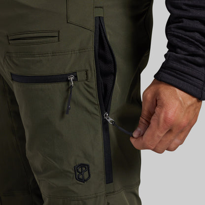 Close-up of olive green outdoor pants with a hand opening a black zipper pocket, showing the fabric texture and a small shield logo near the lower leg.