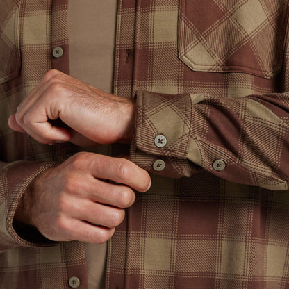 Close-up of a man's hands buttoning the cuffs of a brown and tan plaid flannel shirt over a tan undershirt.