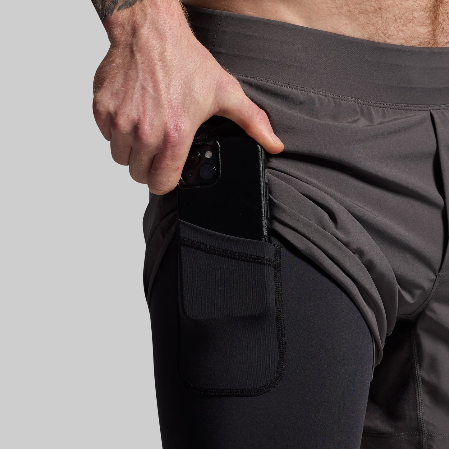 A person wearing gray athletic shorts with a compression section on the left thigh and a mobile phone in their hand.