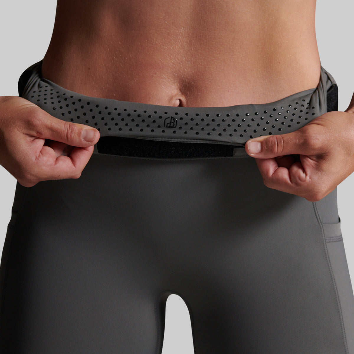 Close-up of a person wearing dark gray athletic leggings and pulling up a matching waistband with both hands. The waistband has elastic grip