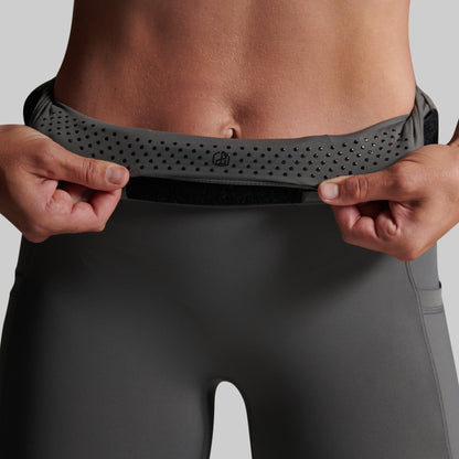 Close-up of a person wearing dark gray athletic leggings and pulling up a matching waistband with both hands. The waistband has elastic grip