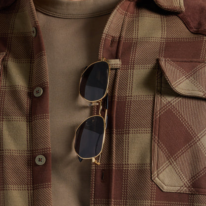 Close-up of a person wearing a brown and tan plaid flannel shirt over a taupe t-shirt, with gold-framed sunglasses hanging from the placket.