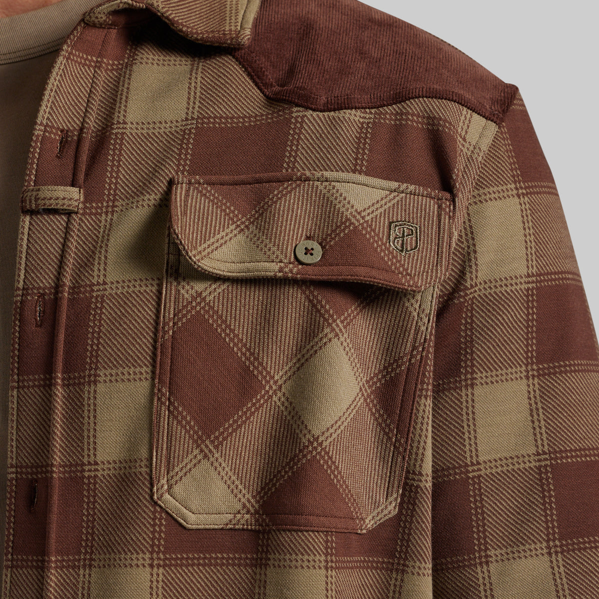 Close-up of a brown and beige plaid flannel shirt, showing a chest pocket with a button flap and a small shield logo patch.