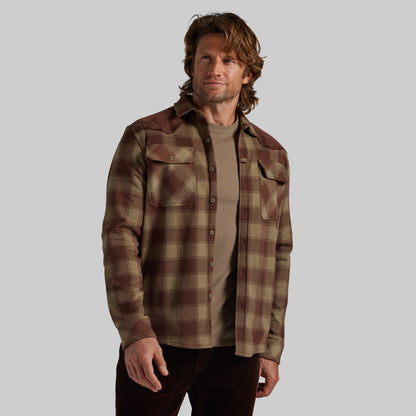 A man wearing a brown and beige plaid button-up flannel shirt over a tan t-shirt, photographed against a solid gray studio background.