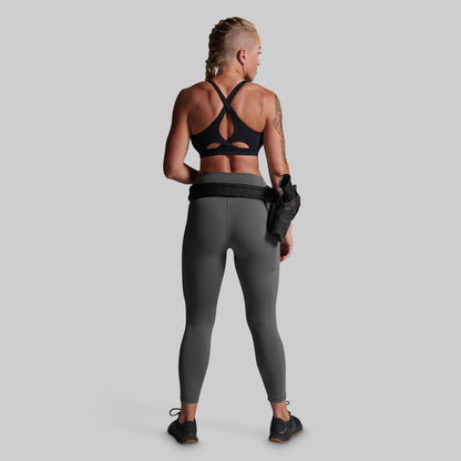 A woman shown from the back wearing dark gray full-length leggings and a black sports bra, standing against a light gray studio background. She is wearing a black belt with a holster