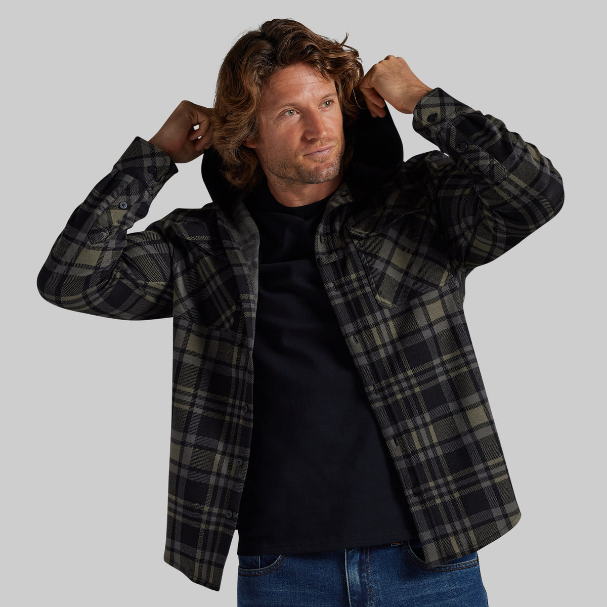 A man wearing a black and olive green plaid hooded overshirt over a black t-shirt and blue jeans, posing with his hands in his hair against a solid gray background.