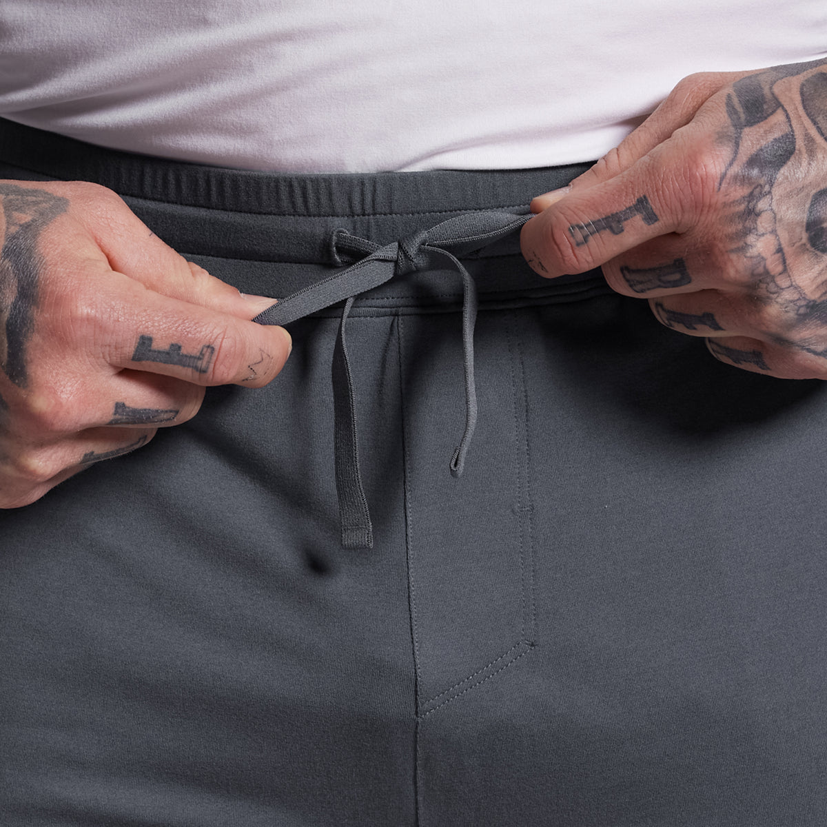 Person adjusting the drawstring of gray shorts with a tattooed hand.