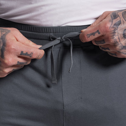 Person adjusting the drawstring of gray shorts with a tattooed hand.