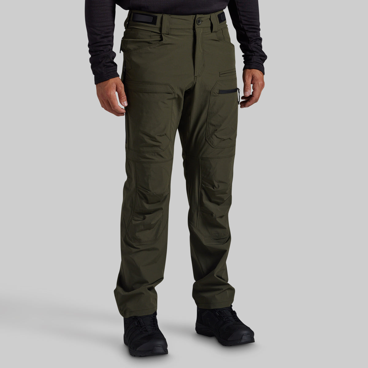 A person wearing olive green outdoor cargo pants with a black long-sleeve shirt and black boots, shown from the waist down against a light gray studio background.