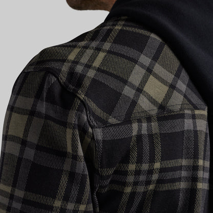 Close-up of a person wearing a dark plaid overshirt or jacket with a black hood, showing the shoulder and upper chest against a light gray background.