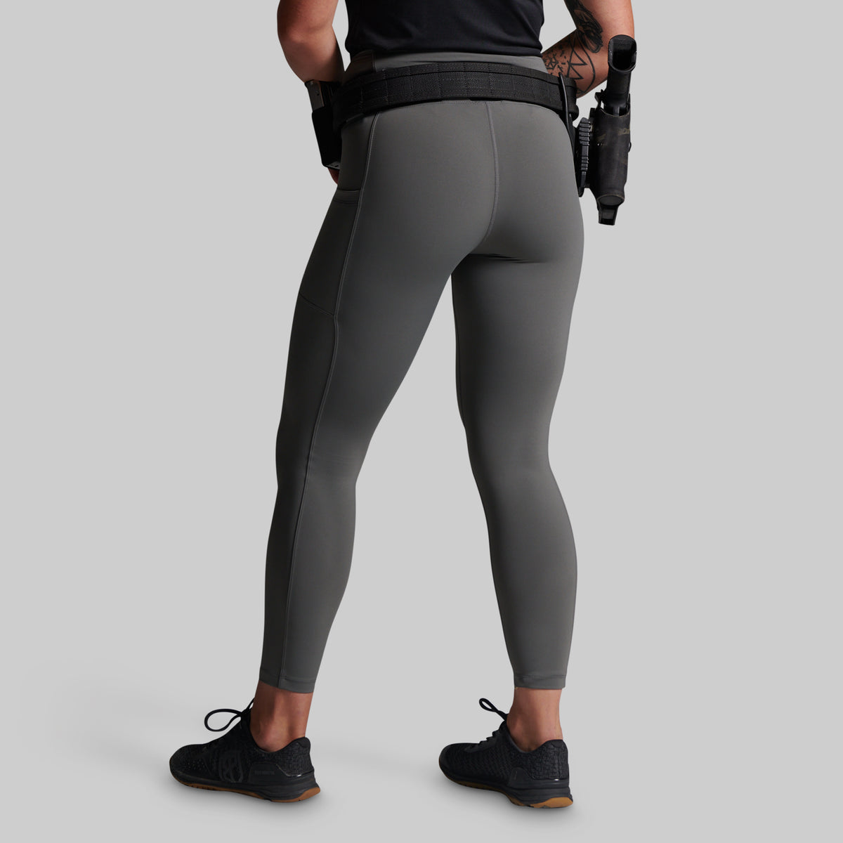 Rear view of a model wearing dark gray full-length athletic leggings with a black waist belt and black sneakers, standing against a solid light gray background.