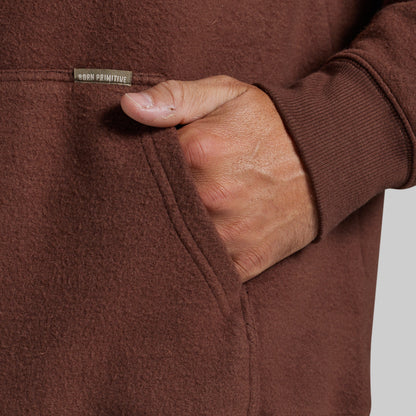 Close-up of a person wearing a brown fleece hoodie, showing a hand in the pocket with a small 'Born Primitive' brand tag visible on the chest.