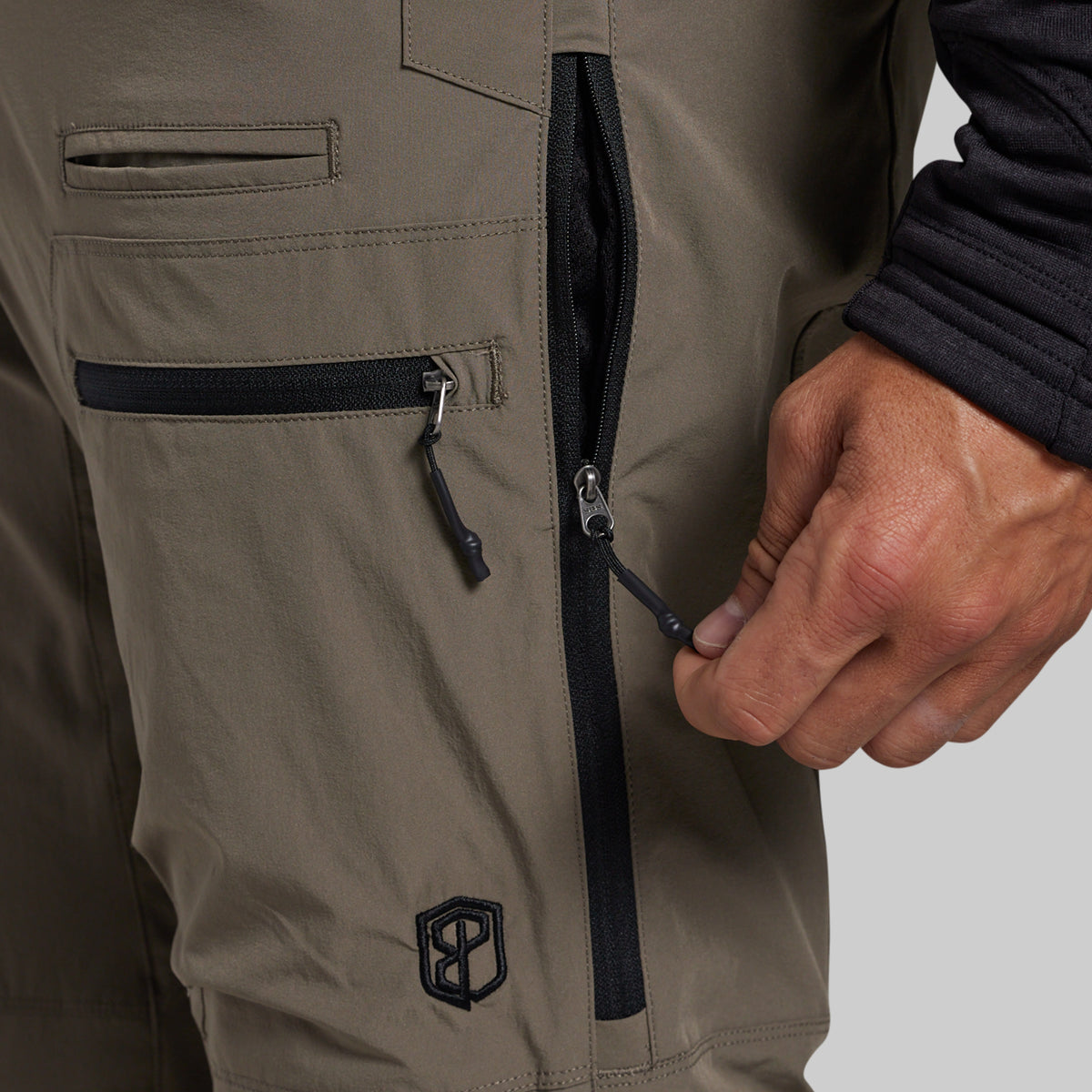 Close-up of a person's hand zipping a black vertical pocket on grey technical pants, showing the fabric texture and a black embroidered logo near the lower leg.