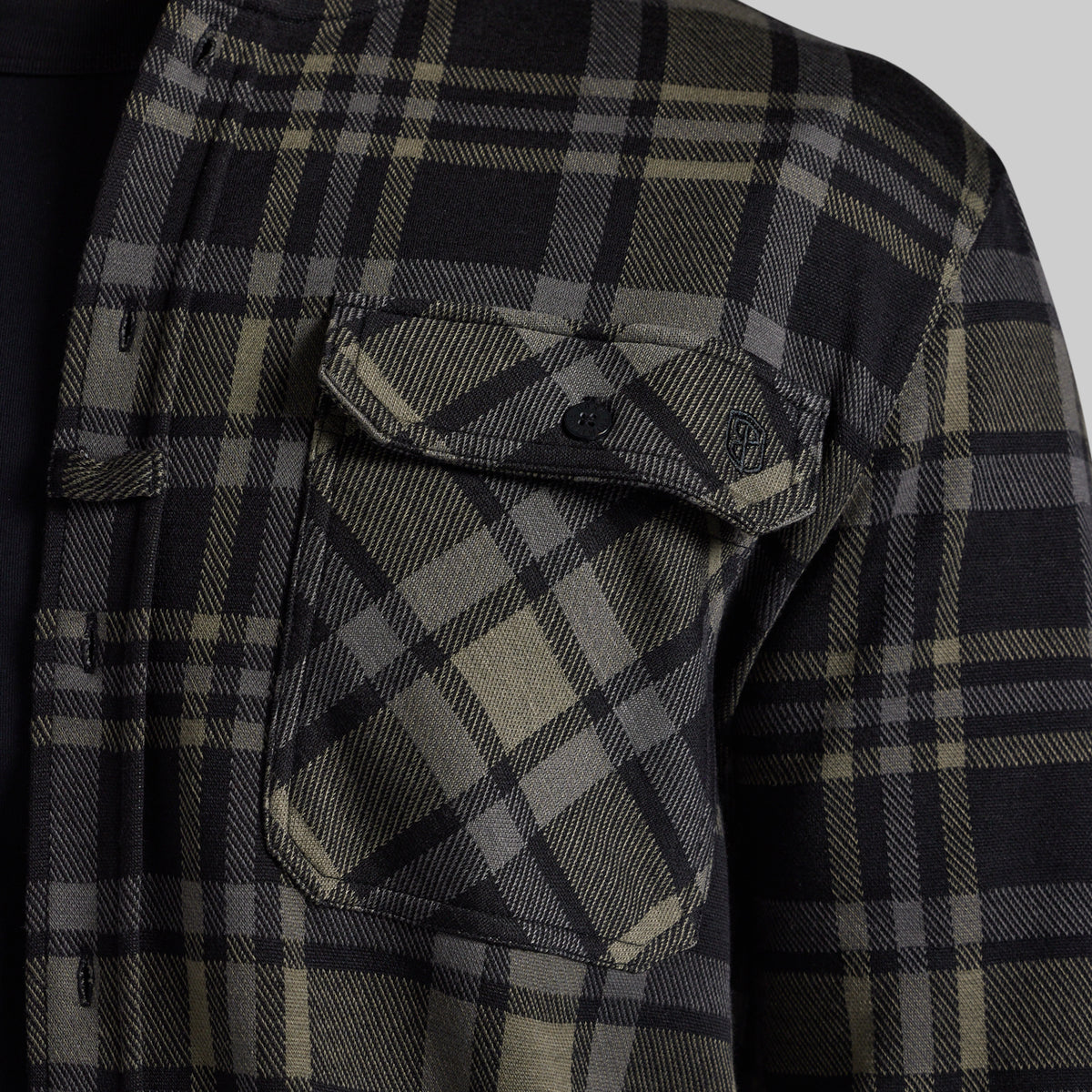 Close-up of a black and greu plaid flannel shirt, showing a chest pocket with a button flap and a small shield logo patch.