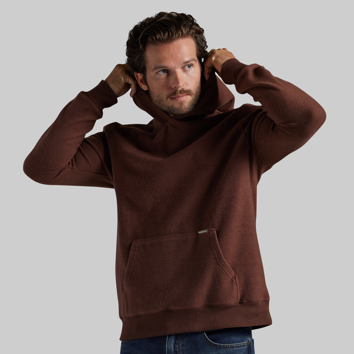 A man wearing a brown fleece pullover hoodie with a kangaroo pocket, shown from the waist up against a solid gray background.