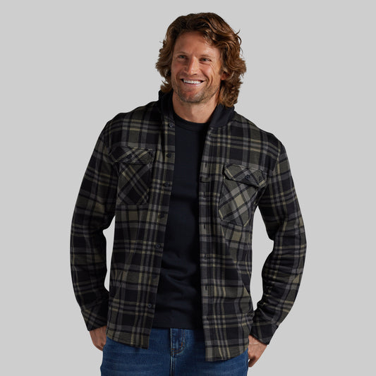 A man wearing a black and green plaid button-up flannel shirt over a black t-shirt, photographed against a solid gray studio background.