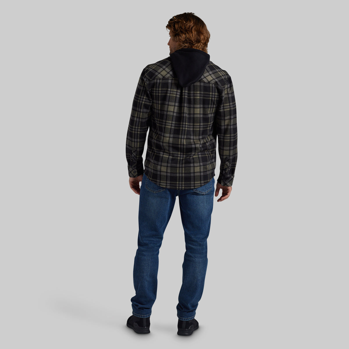 A man viewed from behind wearing a dark green and black plaid hooded jacket, blue jeans, and black shoes, set against a solid gray studio background.