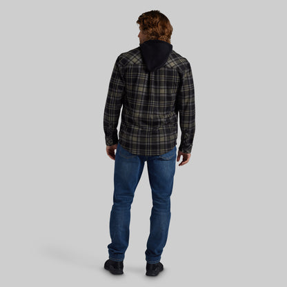 A man viewed from behind wearing a dark green and black plaid hooded jacket, blue jeans, and black shoes, set against a solid gray studio background.