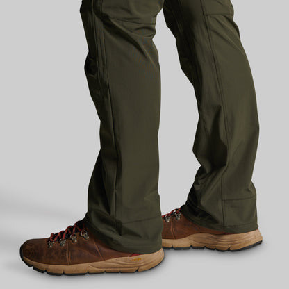 Close-up of a person's legs wearing olive green pants and brown hiking boots against a plain light gray studio background.