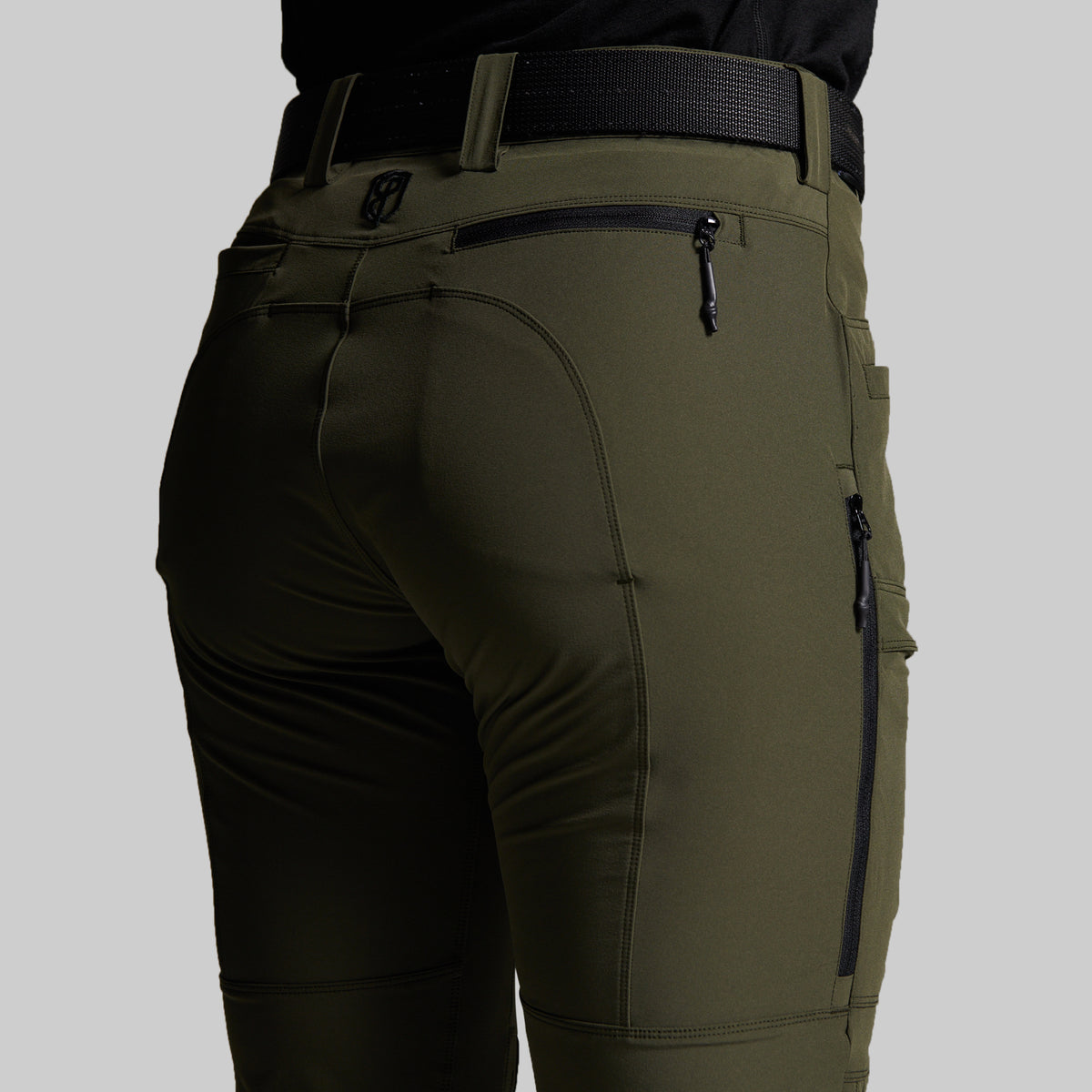 Close-up of a person wearing olive green outdoor pants with a black belt, showing the back pocket and zippered details.