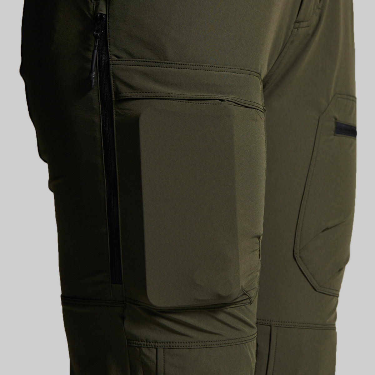 Close-up of olive green technical outdoor pants showing a large front flap pocket, side zippered pocket, and angled cargo pocket on the thigh.