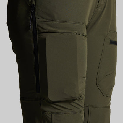 Close-up of olive green technical outdoor pants showing a large front flap pocket, side zippered pocket, and angled cargo pocket on the thigh.