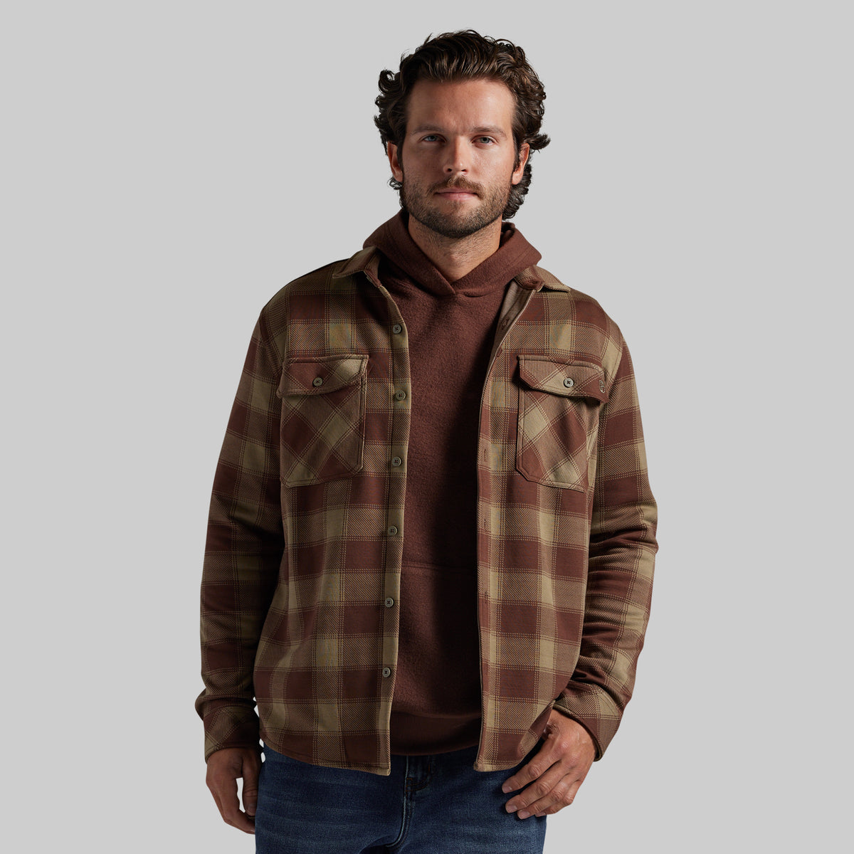 A man wearing a brown and tan plaid overshirt over a brown hoodie and blue jeans, standing against a solid gray background.