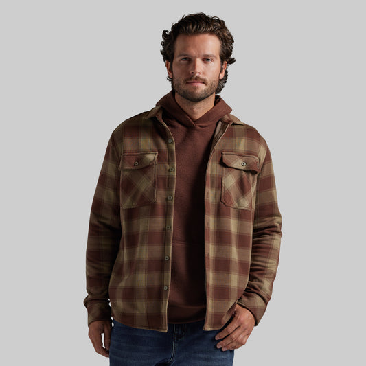 A man wearing a brown and tan plaid overshirt over a brown hoodie and blue jeans, standing against a solid gray background.
