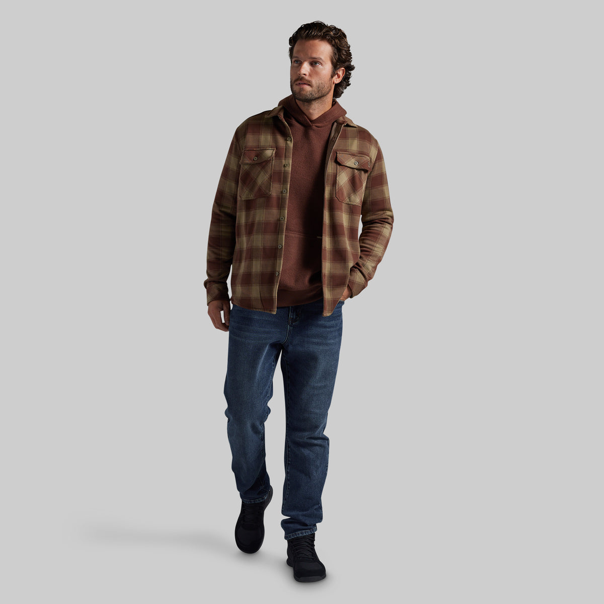 A man modeling a brown and tan plaid flannel shirt jacket over a brown hoodie, paired with blue jeans and black boots, against a solid gray background.