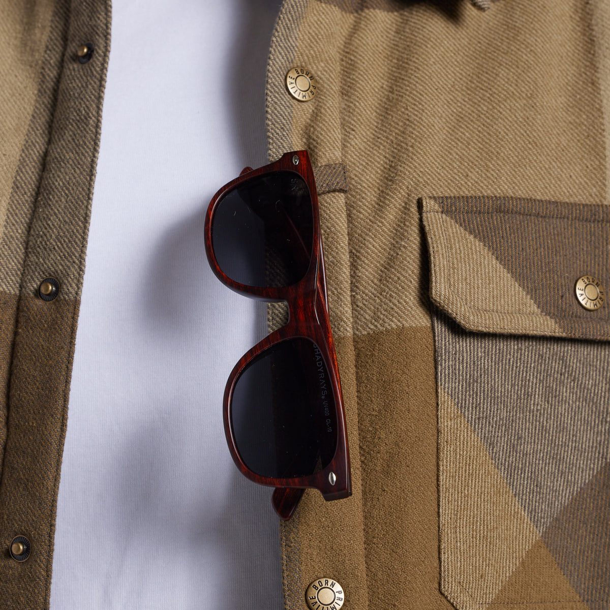 A pair of sunglasses hanging from the sunglass loop of a tan and brown plaid flannel shirt, worn over a white t-shirt.