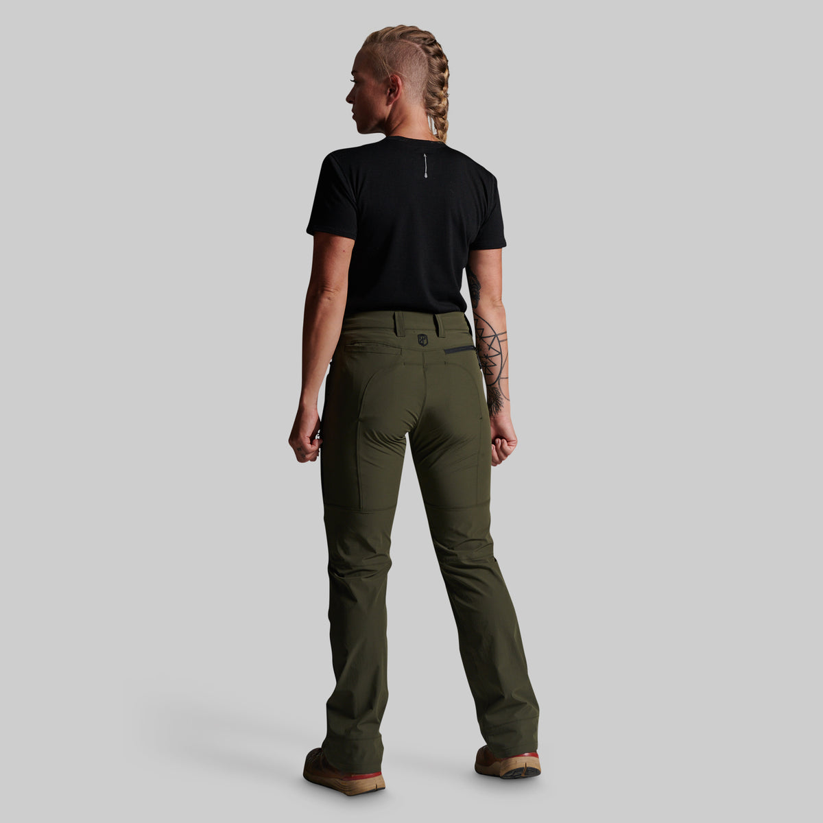 A woman shown from the back wearing olive green outdoor pants, a black t-shirt, and brown boots against a solid gray studio background.