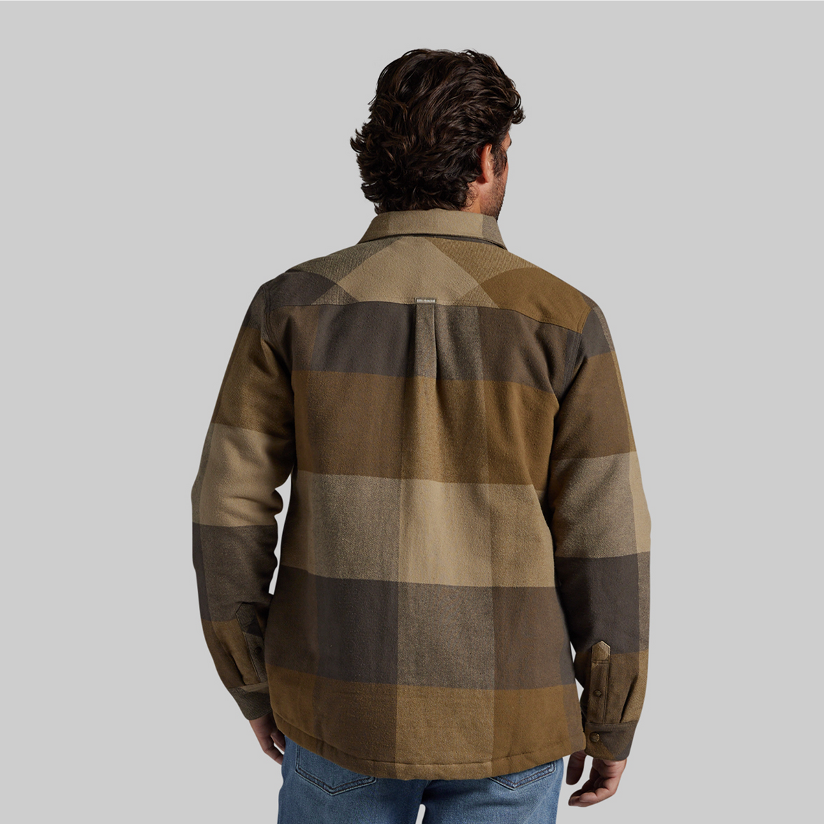 A man viewed from behind wearing a long-sleeve plaid overshirt in earthy brown, tan, and olive tones, paired with blue jeans, against a solid gray studio background.