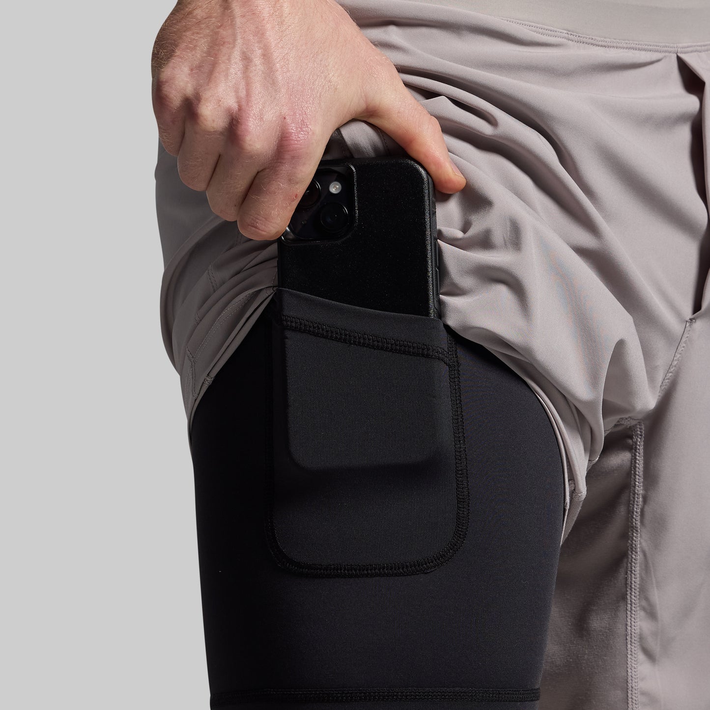 A person wearing a pair of grey athletic shorts with a black compression liner and a phone pocket on the right thigh.